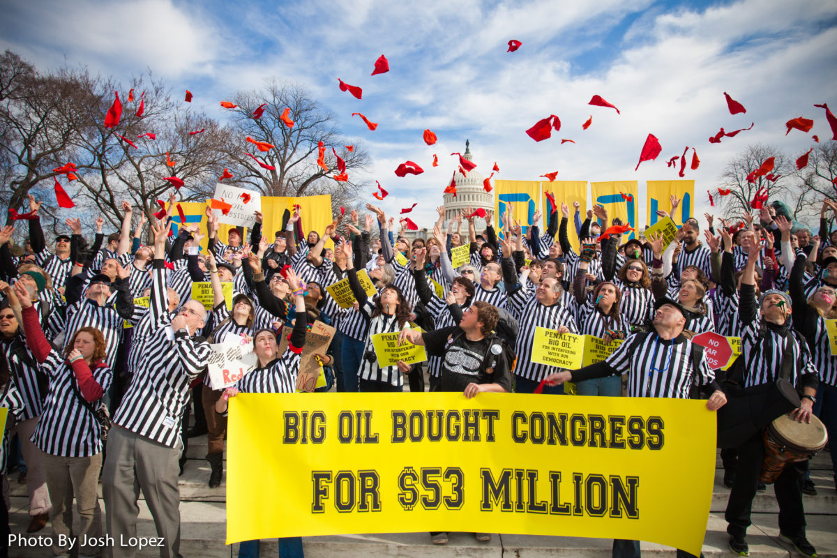 Protestors outside of the US Capitol Building dressed as sports referees throw red flags while holding a sign that reads "Big Oil Bought Congress for $53 Million"