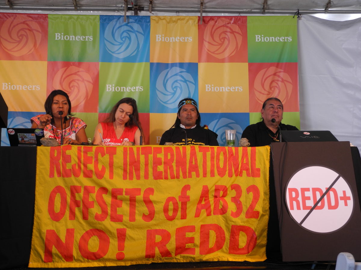 Four speakers sitting at a table behind a banner that says "Reject international offsets of AB32 No! Redd"
