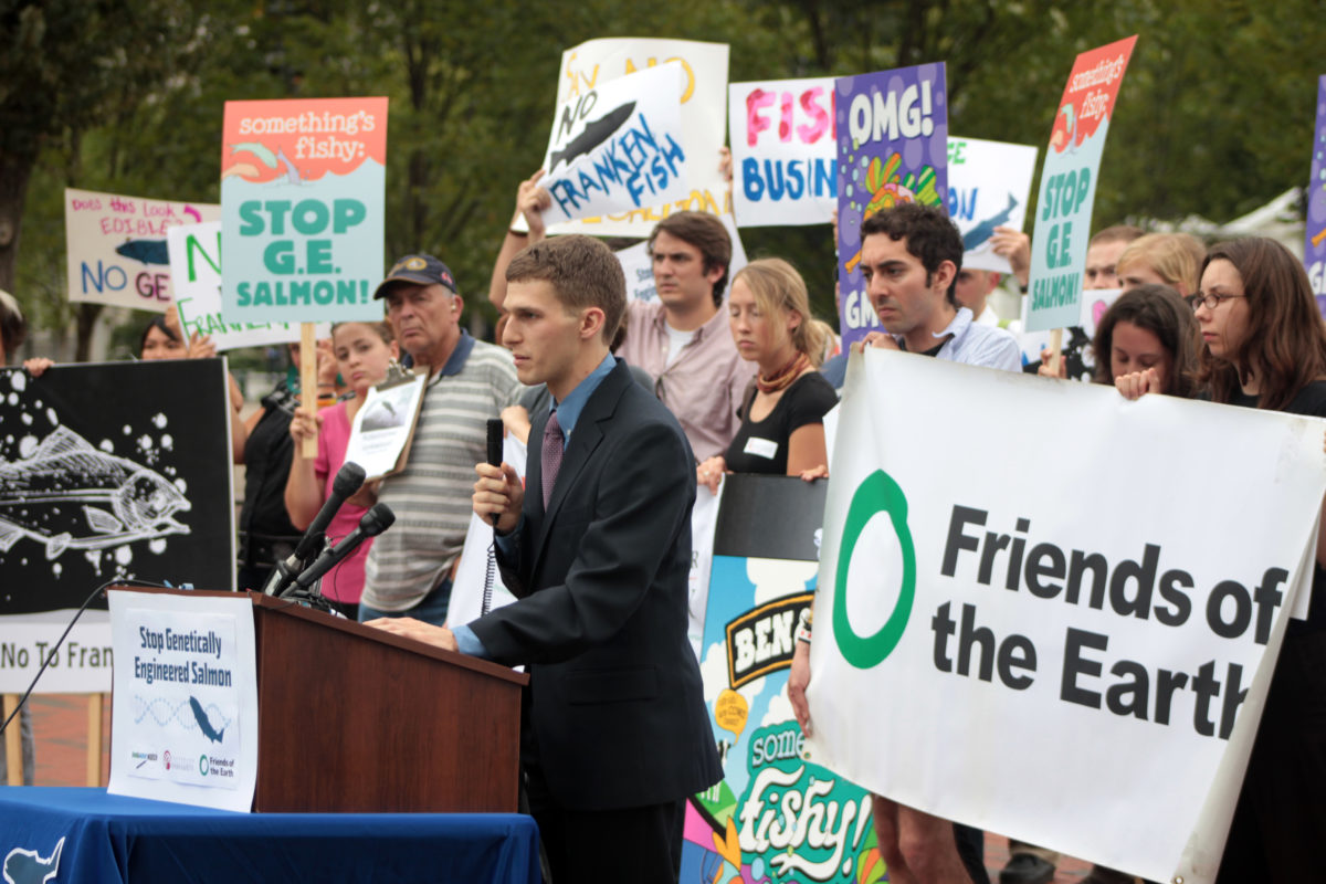 Protest speaker standing at a podium in front of activists holding a Friends of the Earth sign and various signs against genetically engineered salmon