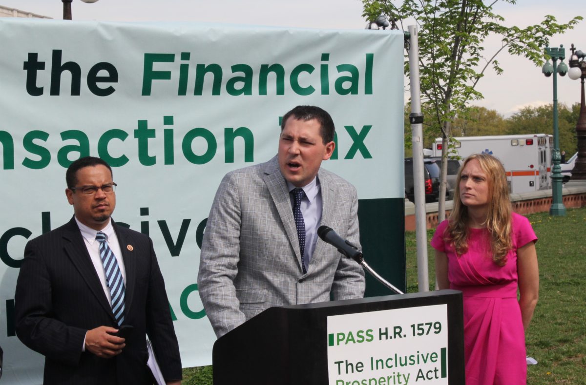 Three speakers stand behind a podium with the sign "Pass H.R. 1579 The Inclusive Prosperity Act"