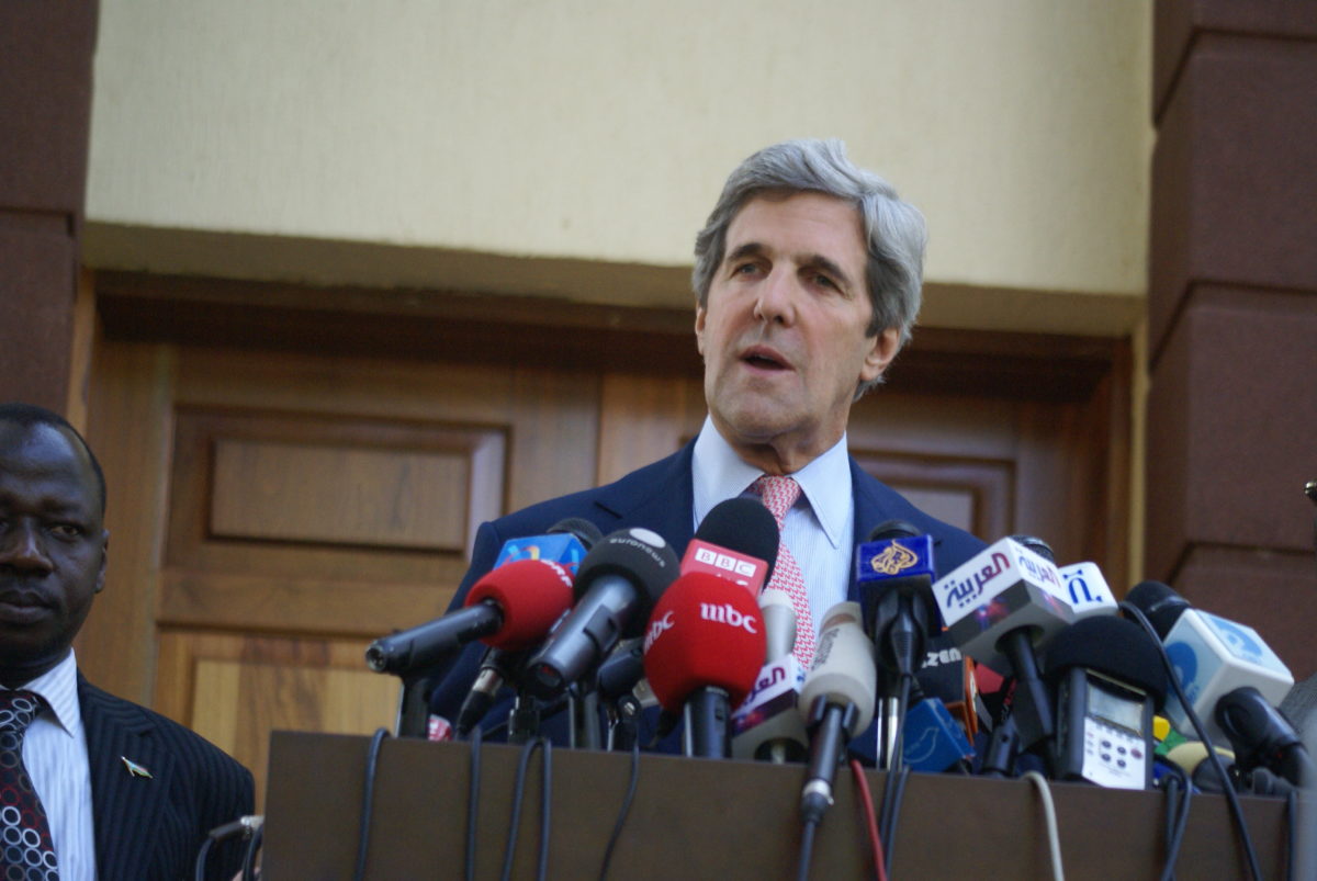 Former Senator John Kerry giving a press conference in front of a bay of microphones