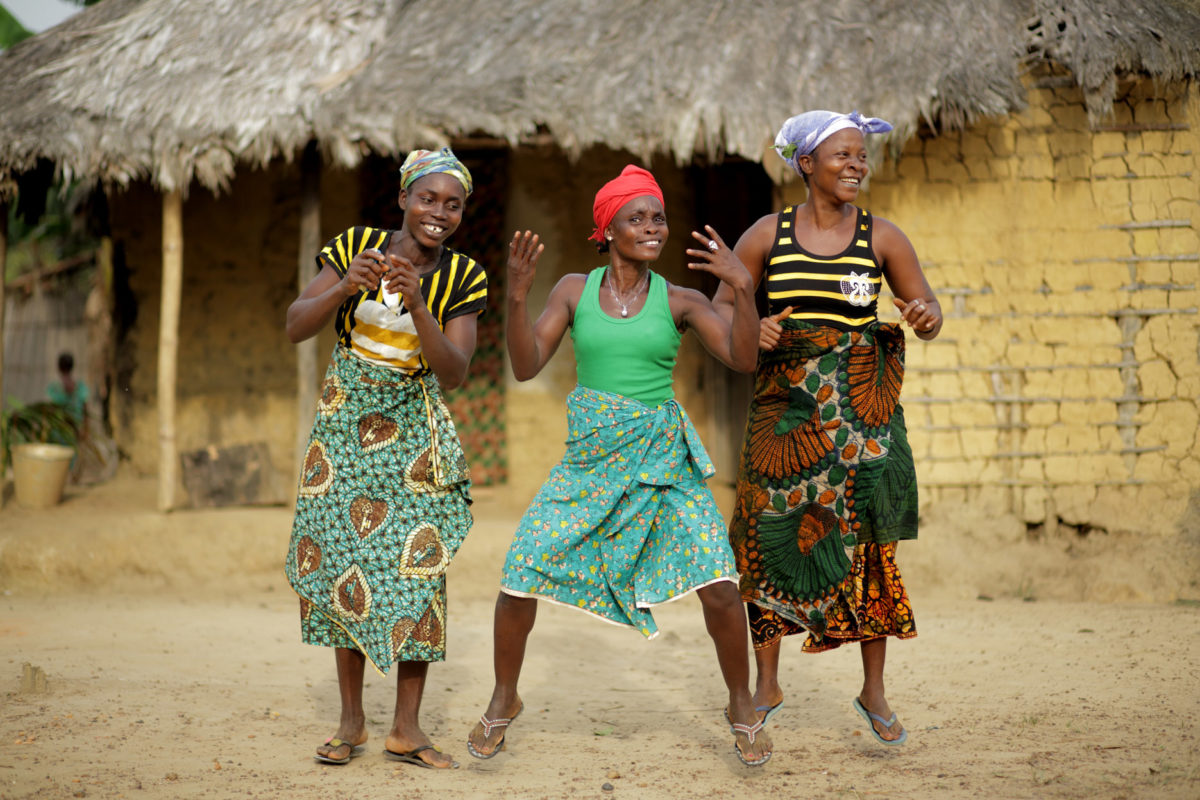 Three African women dancing in front of a house