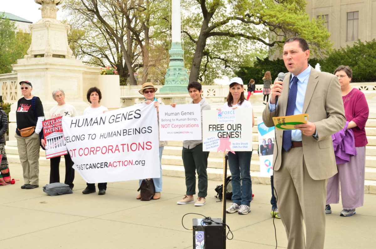 Speaker stands in front of protestors holding signs stating "Human genes belong to human beings not corporations"