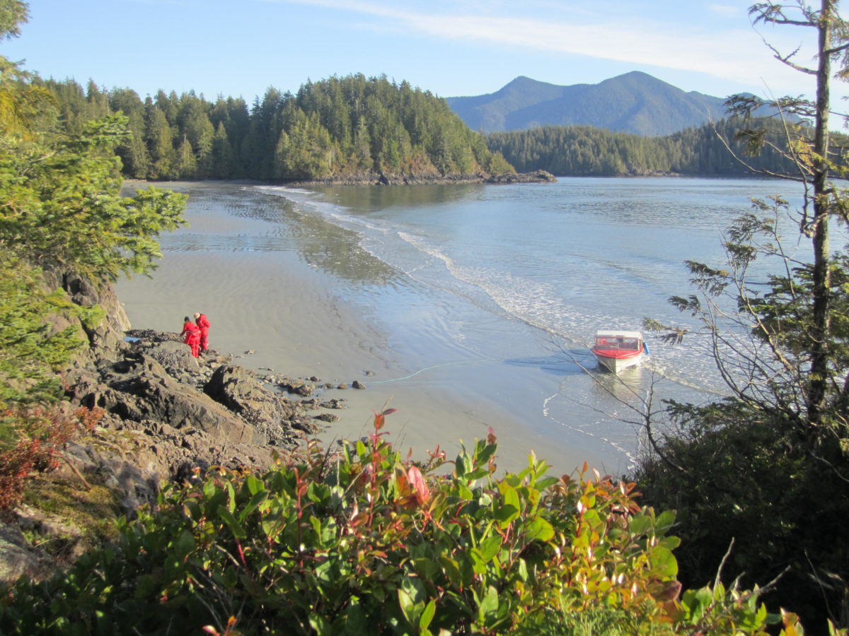 Three people in red standing looking at a mound of sand on the beach with a boat sitting nearby in the water with a forest and mountains in the background