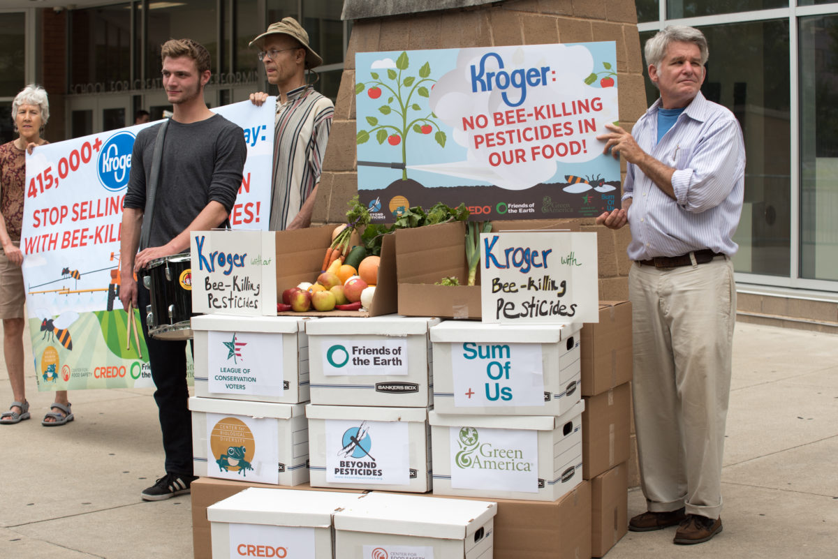 Activists holding signs urging Kroger stores to stop selling food grown with pesticides with a stack of cardboard boxes with environmental group logos on them