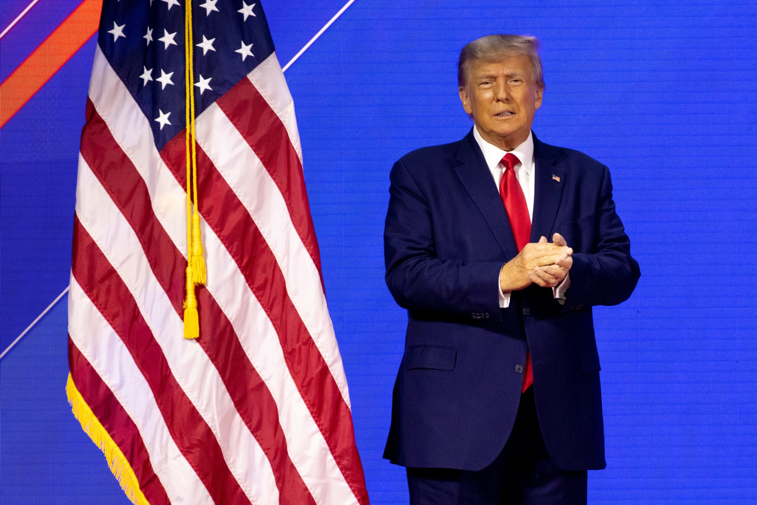 May contain one person in a suit with a red tie standing next to a flag.