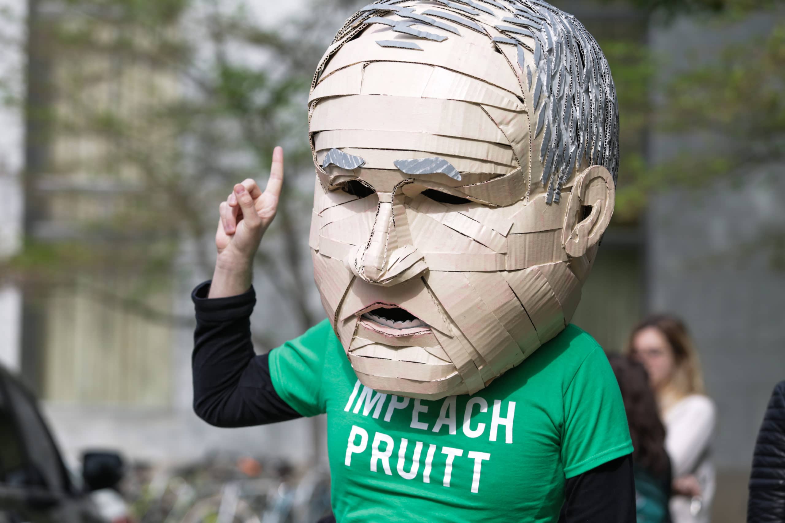 An activist wearing a handcrafted head that resembles Scott Pruitt's wearing a shirt that says "Impeach Pruitt"