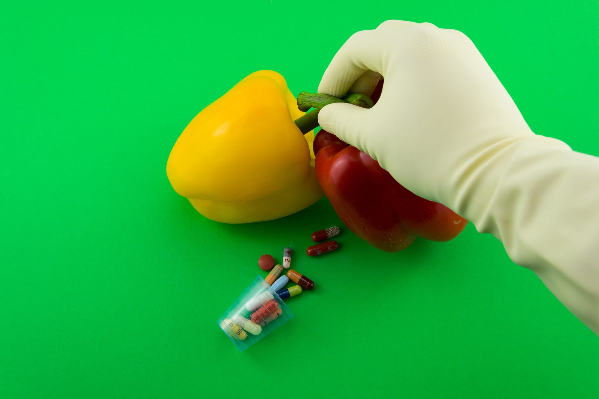 Red and Yellow Bell Peppers Held by Gloved Hand, Next to Spilled Pills