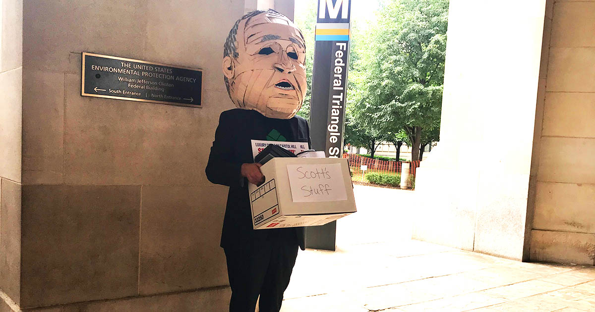 An activist wearing a handcrafted head that resembles Scott Pruitt's holding a cardboard box that says "Scott's Stuff" outside of a DC Metro station