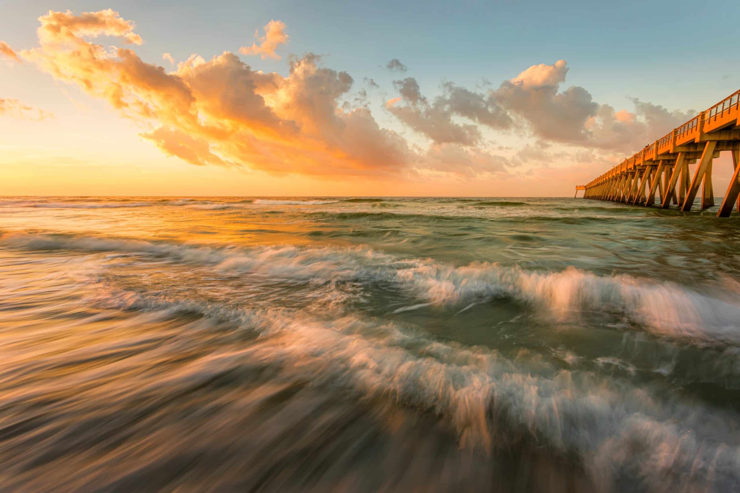 May contain a pier at sunset with orange clouds and waves.