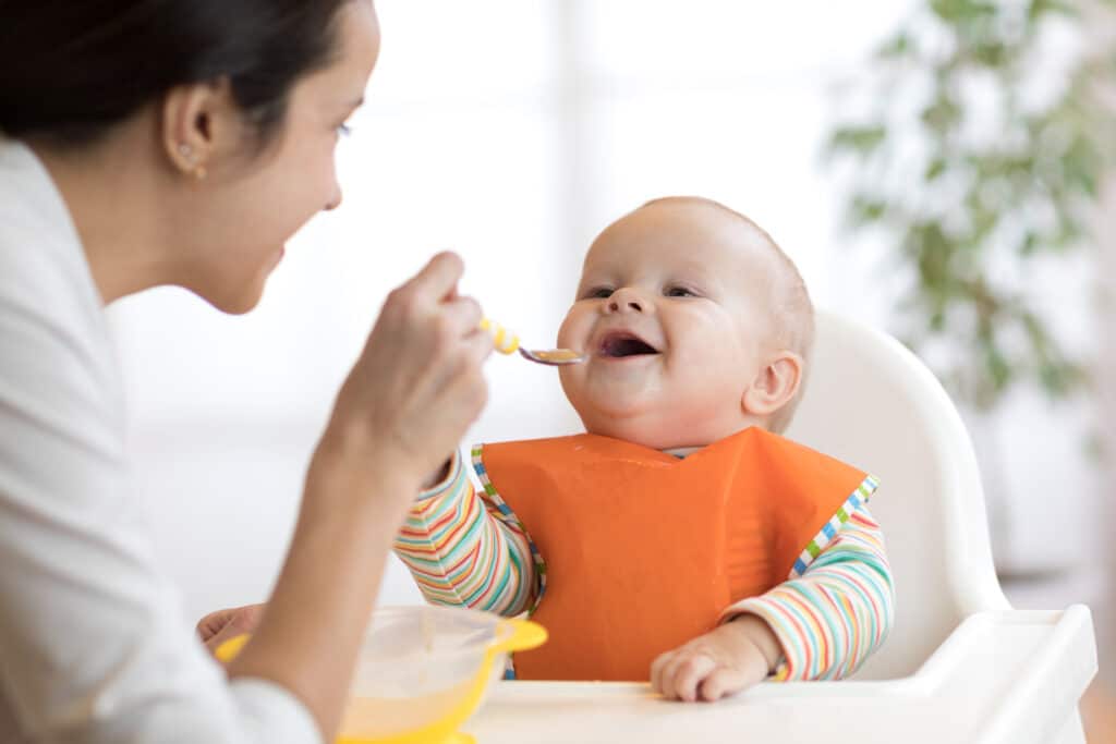 May contain two people, one feeding a baby in a high chair.