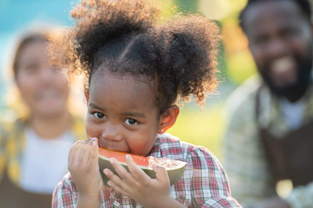 May contain a child eating watermelon with two people in the background.