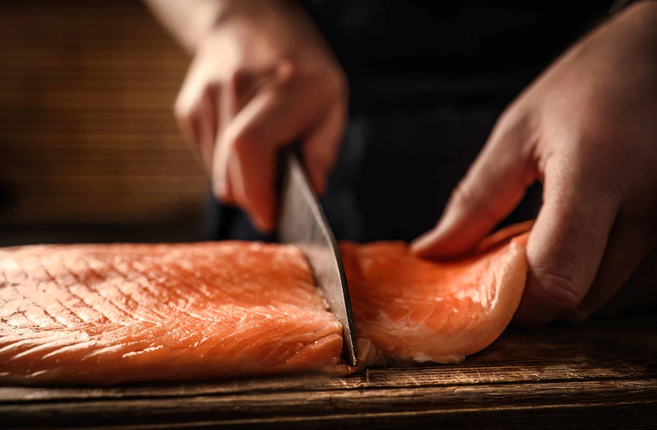 May contain a person's hands slicing a raw salmon fillet on a wooden board.
