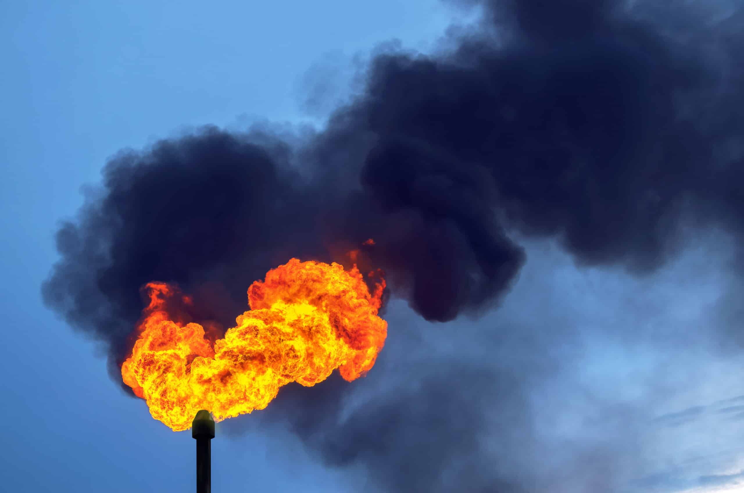 May contain a flare stack with bright orange flames and dark smoke against a blue sky.