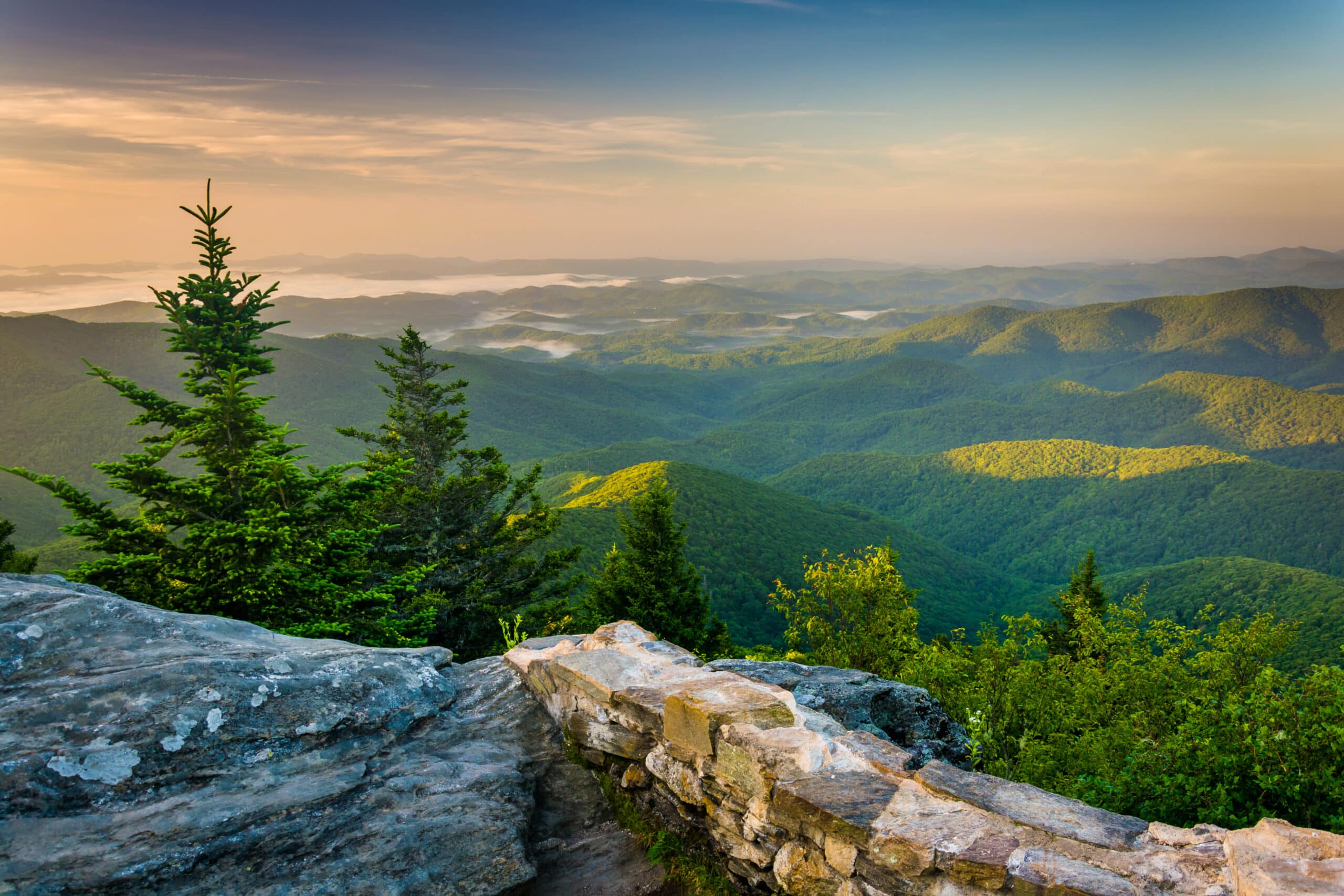 May contain a rocky overlook with a stone wall and evergreen trees facing rolling green hills at sunrise.