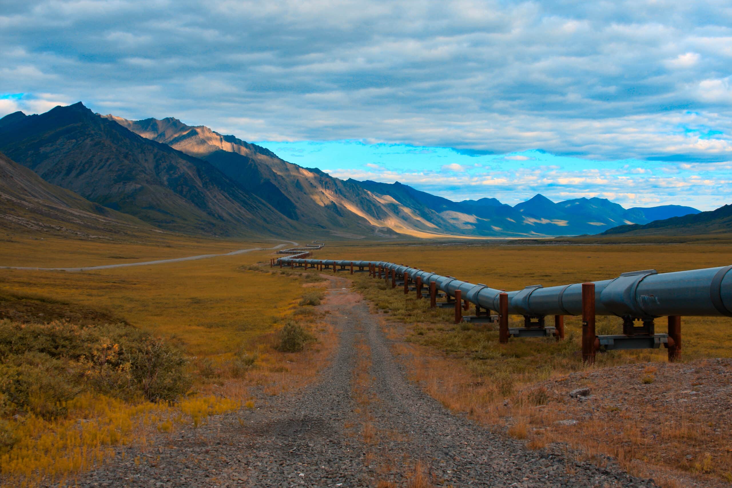 May contain pipeline stretching across a vast, golden, grassy plain toward distant mountains under a cloudy sky.